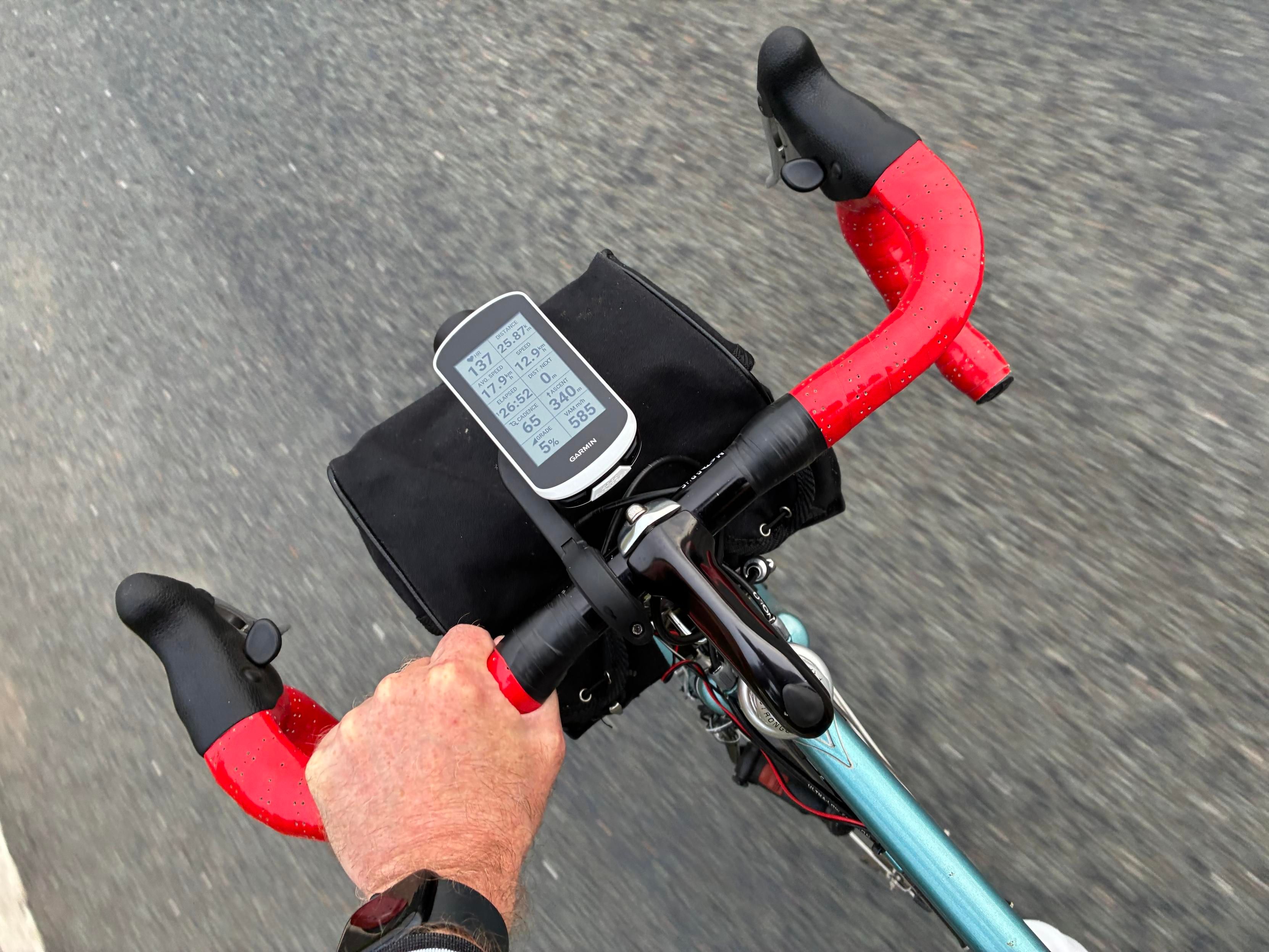Close-up view of bike handlebars with red tape, a cycling computer displaying various metrics, and a black bag on the bike. 