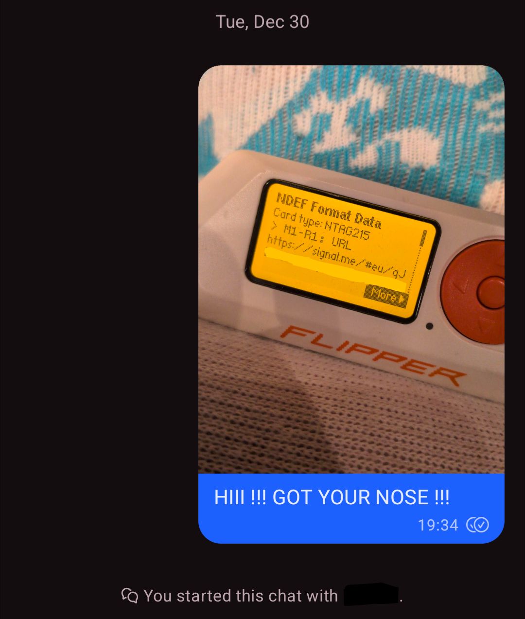 Signal message, beginning a chat on 2025-12-30 19:34. it's an image of a flipper zero showing some NTAG-215 data, which is a URL containing a Signal profile link. behind the flipper zero are some blue striped though highs with a cat face. 

attached to the image proving possession of the NFC data is a message: "HIII !!! GOT YOUR NOSE !!!"