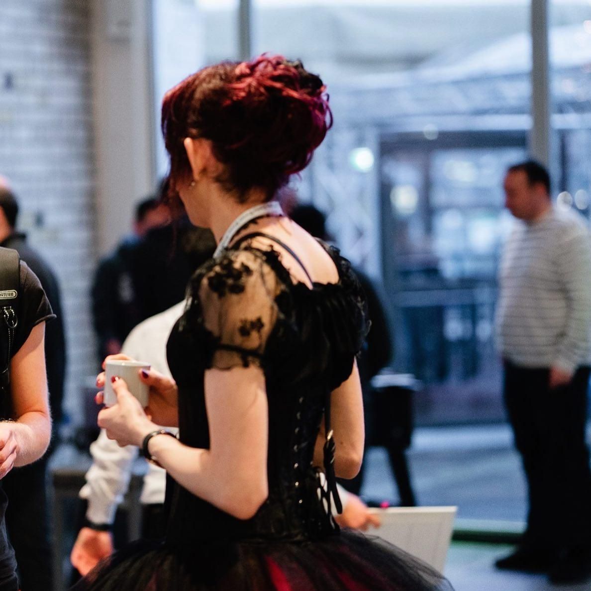 Photo of Emelia at the conference from over the left shoulder. She is holding a coffee cup with both hands, and facing away from the camera. 

Emelia is wearing a black corset, with lace puff sleeves, her hair is black and up in a fancy updo with a mixture of dark red and magenta highlights. At the bottom of the photo you can see the tulle skirt of her ballgown peaking through with pops of reds and pinks through black.

Behind Emelia is a man standing in business casual, his face blurred.

This photo was taken by the React Day Berlin 2019 event photographers (unfortunately uncredited by name).