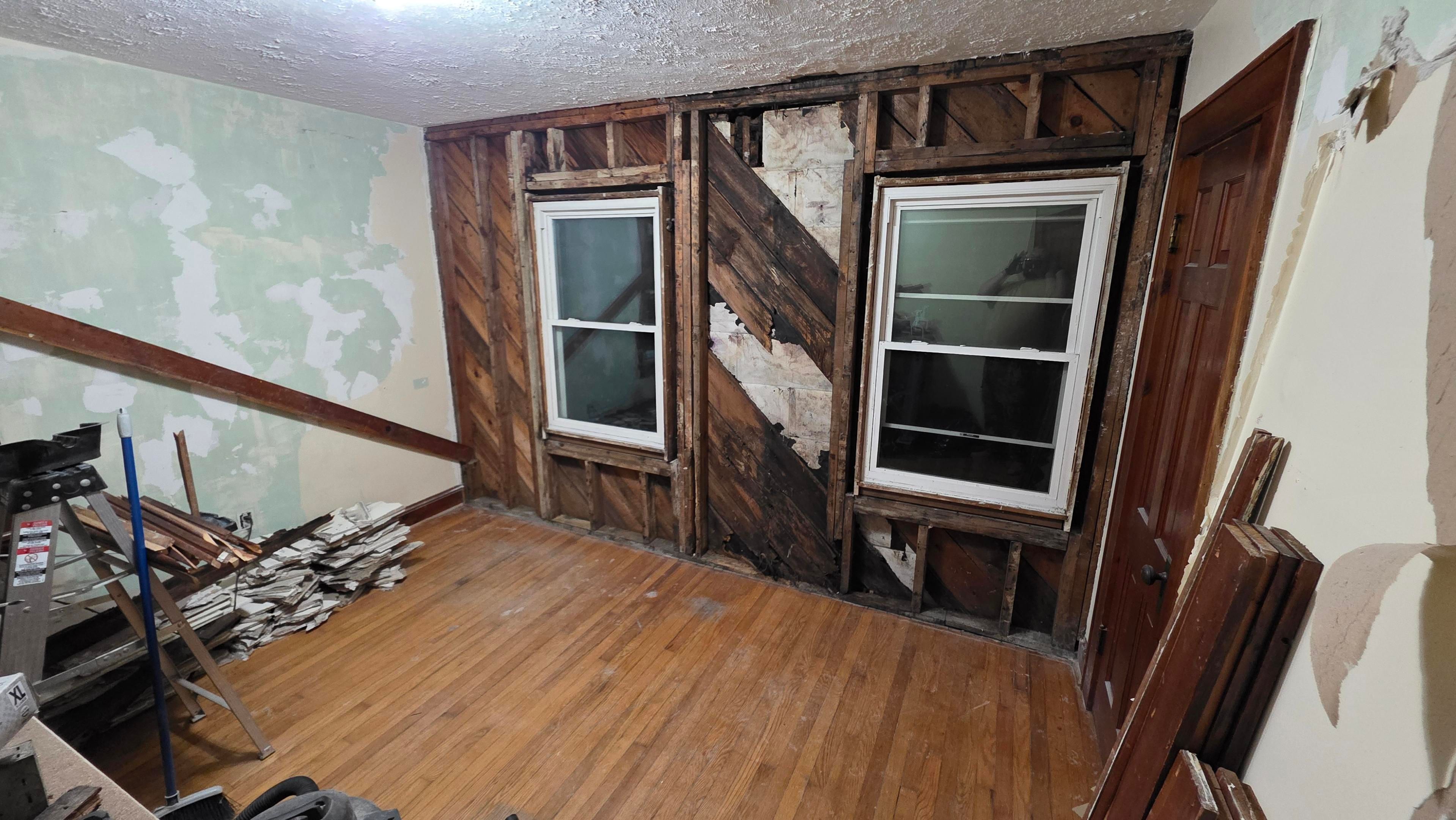 A demolished wall with two windows, rotted in the middle. Swept hardwood floor and stacked chunks of plaster.