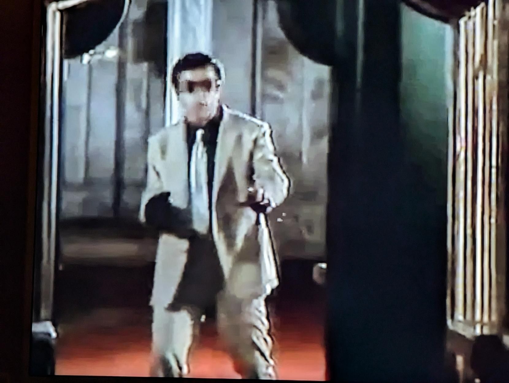 Screenshot from a movie. A man in a pale suit, black shirt and white tie. He is wearing dark sunglasses. His right hand is encased in a black leather glove with stiff fingers. He's obviously a wrong'un