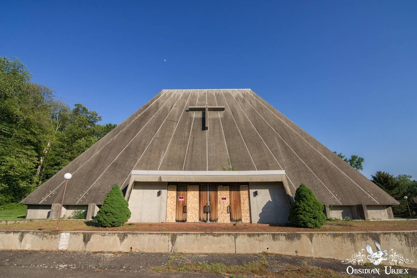 This is an image of a uniquely designed, abandoned church. The building is pyramid-shaped with a cross on the front and boarded-up doors.