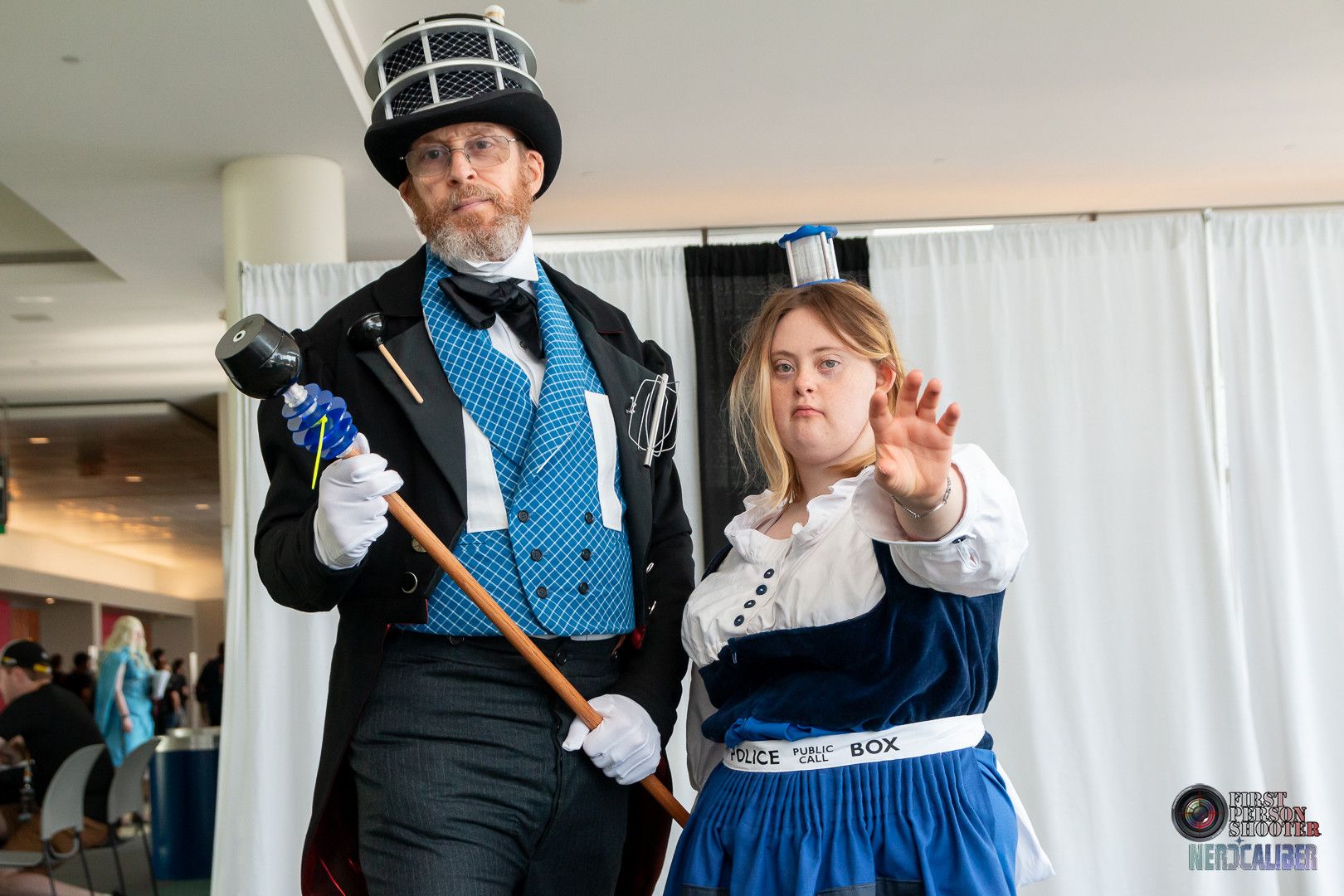 A man cosplaying as a Dapper Dalek wearing a full suit, tailcoat and top hat next to a young woman with Down syndrome cosplaying as a TARDIS