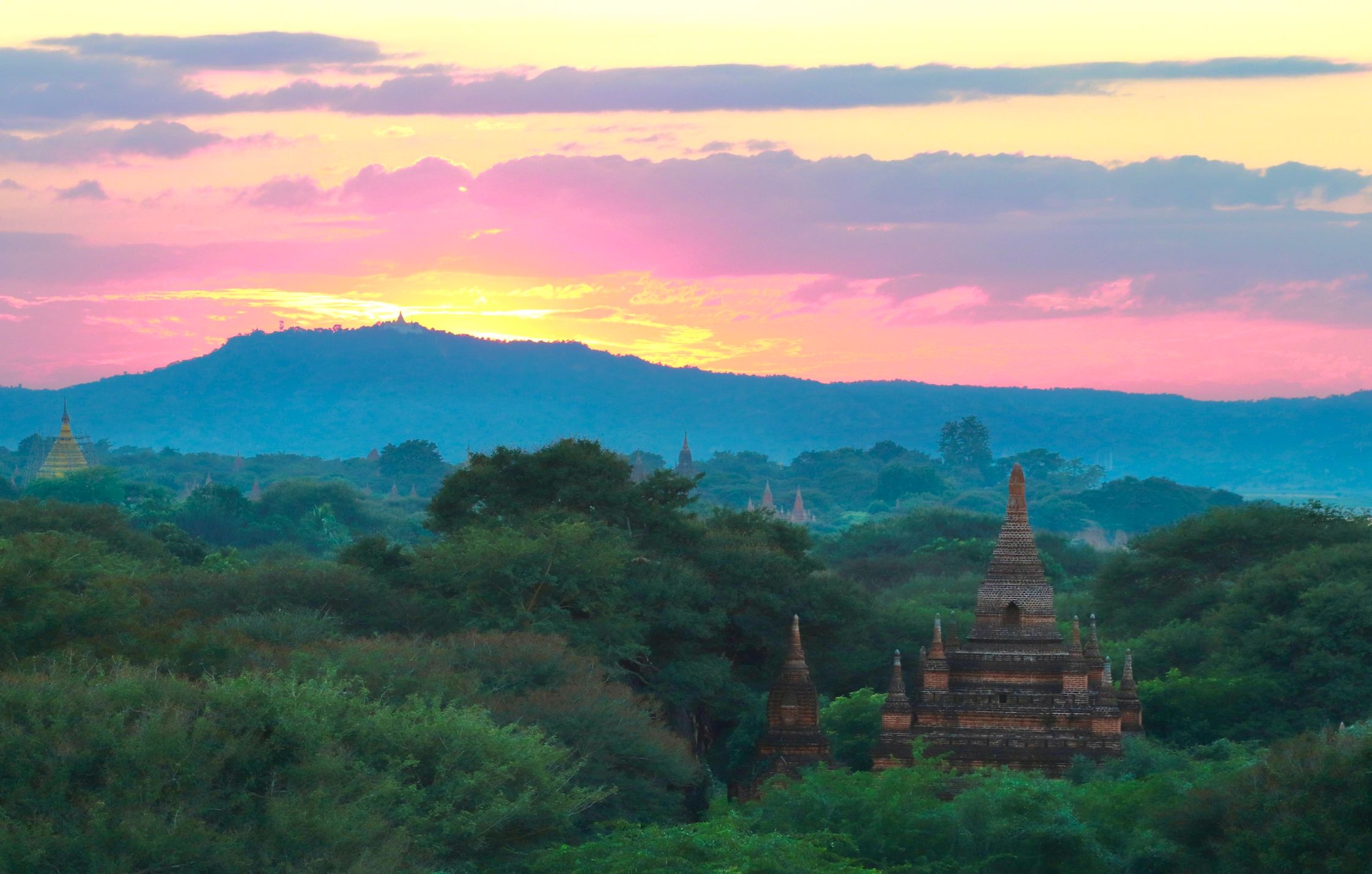 the sunset creates a veritable rainbow in the sky that goes from yellow to pink to lavender to blue to a sort of greenish white. on the ground, lush jungle surrounds a few scattered temples, as a small mountain/large hill rises in the distance
