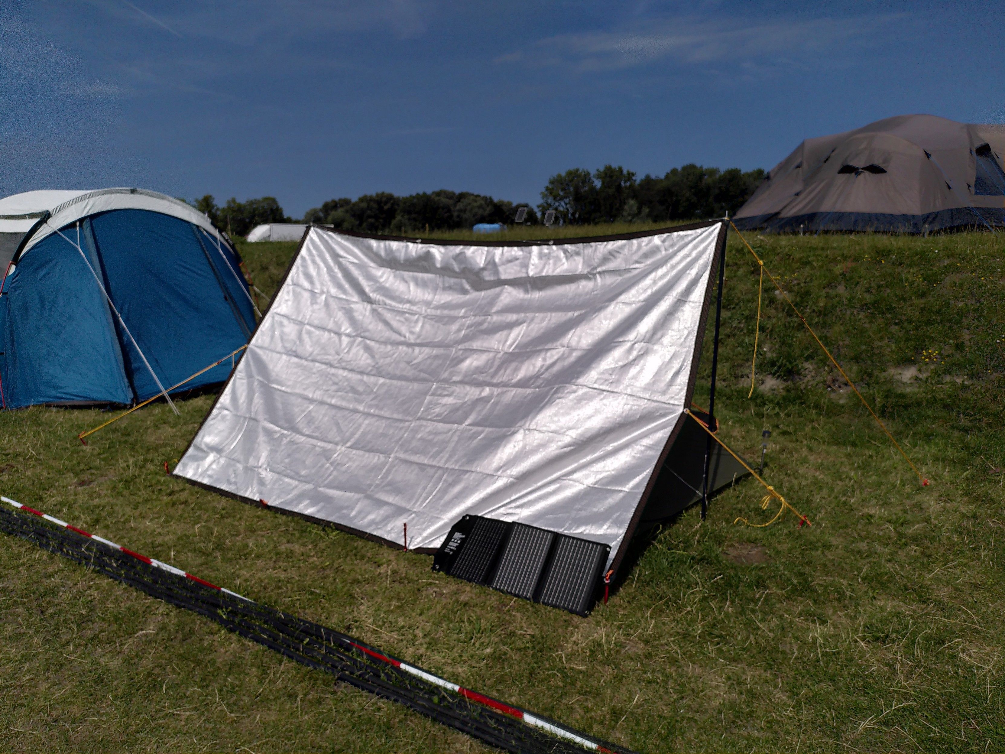 A shiny silver coloured tarp pitches as a lean-to setup. In the bottom right corner is a small solar panel. The tarp is held up with yellow guy lines. There's a tent next to the tarp.