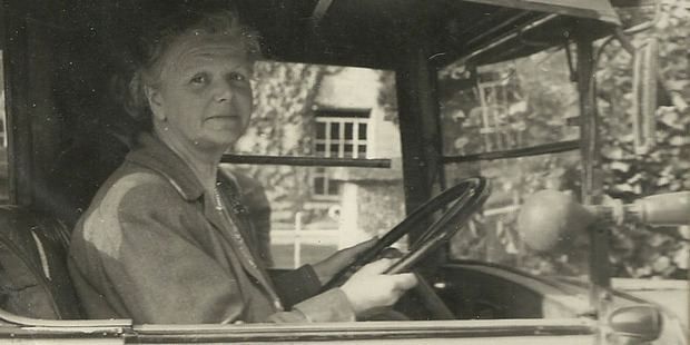 Black and wihte photo . British composer Dorothy Gertrude Howell looking into the camera at the left side. Sitting in a car. Hands on the steering-wheel. House and trees visible through the car-windows