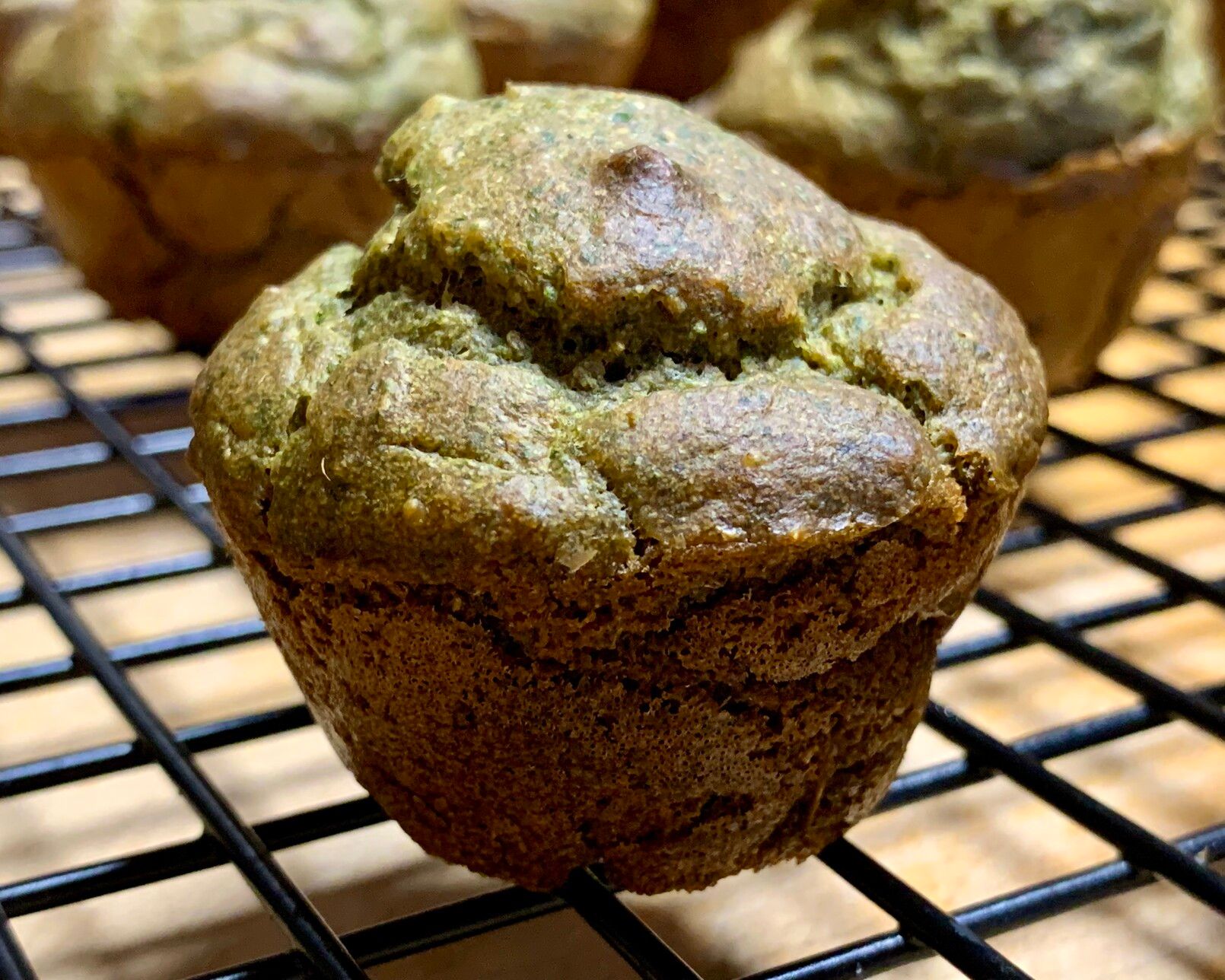 A close up of a small green fluffy muffin on a black rack with more muffins in the background. The outsides of the muffin are slightly browned from baking but is cracked to reveal verdant fluffy insides. 