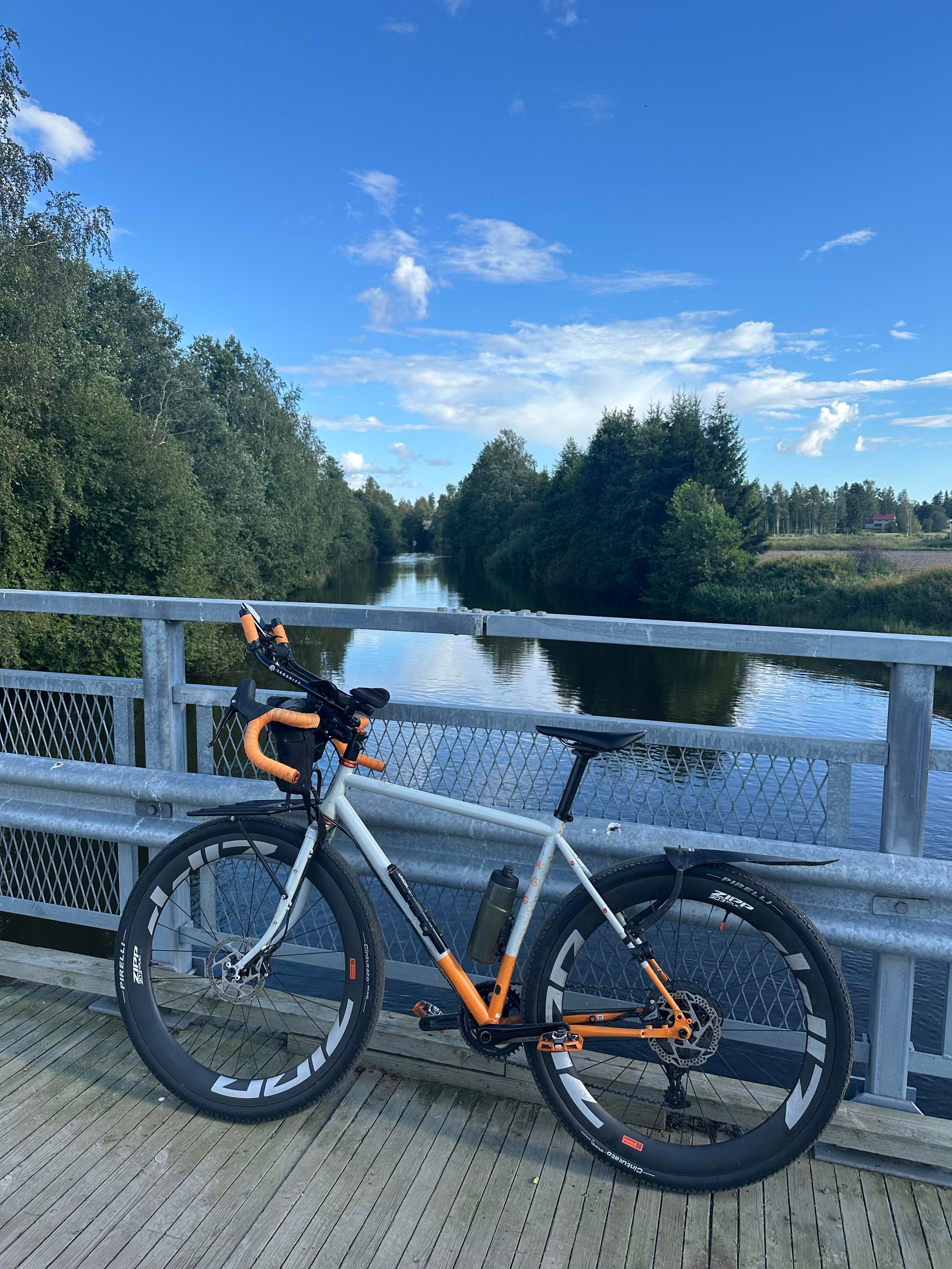 Harmaa/oranssi pyörä nojaa joen ylittävän sillan kaiteeseen. Taustalla joki ja vihreää metsää. Taivas on sininen, paitsi valkoisia pilvenhattaroita siellä täällä.