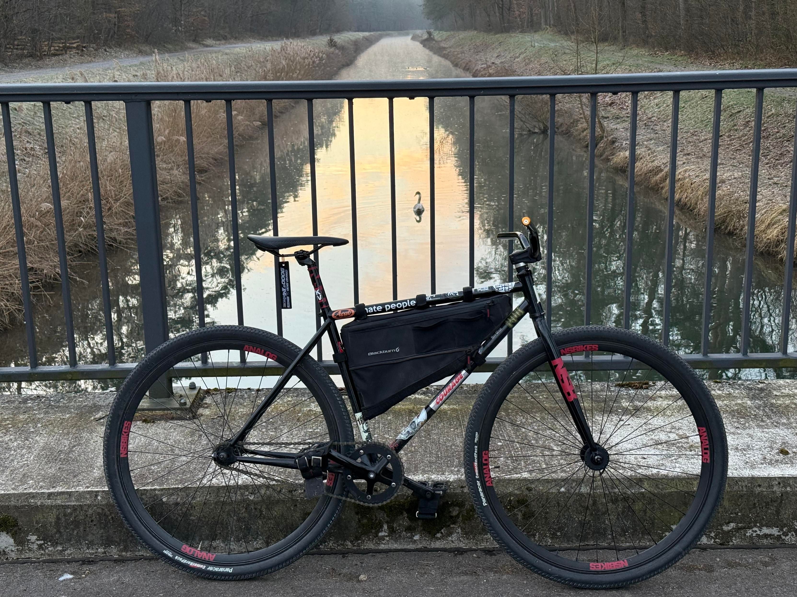A black bicycle is parked on a bridge overlooking a peaceful, reflective waterway surrounded by trees and tall grass. The early morning light casts a soft glow on the scene, with a swan visible in the water.