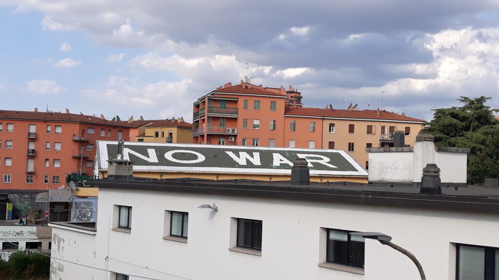 Tetto dell'Ex Mercato di San Donato (Bologna) ripreso dal ponte di S. Donato. Sullo spiovente del tetto è visibile la scritta bianca su fondo nero "No War". Sullo sfondo ci sono dei palazzi e un cielo in parte nuvoloso. 