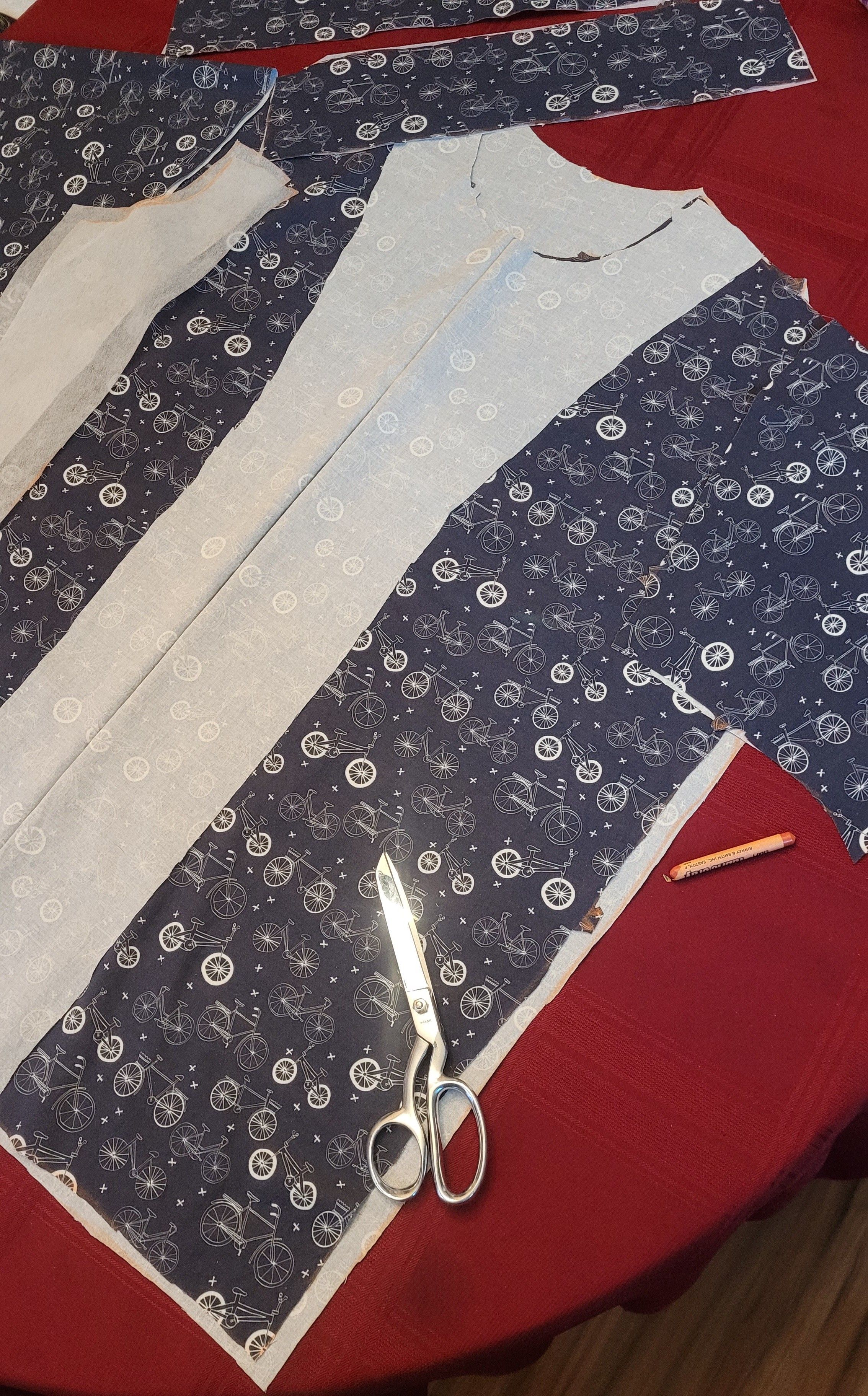 Fabric with bicycles on it in blue neatly cut out on a dining table, Ginger scissors on a red burgundy tablecloth. You can see what looks like wood flooring but it is really just vinyl