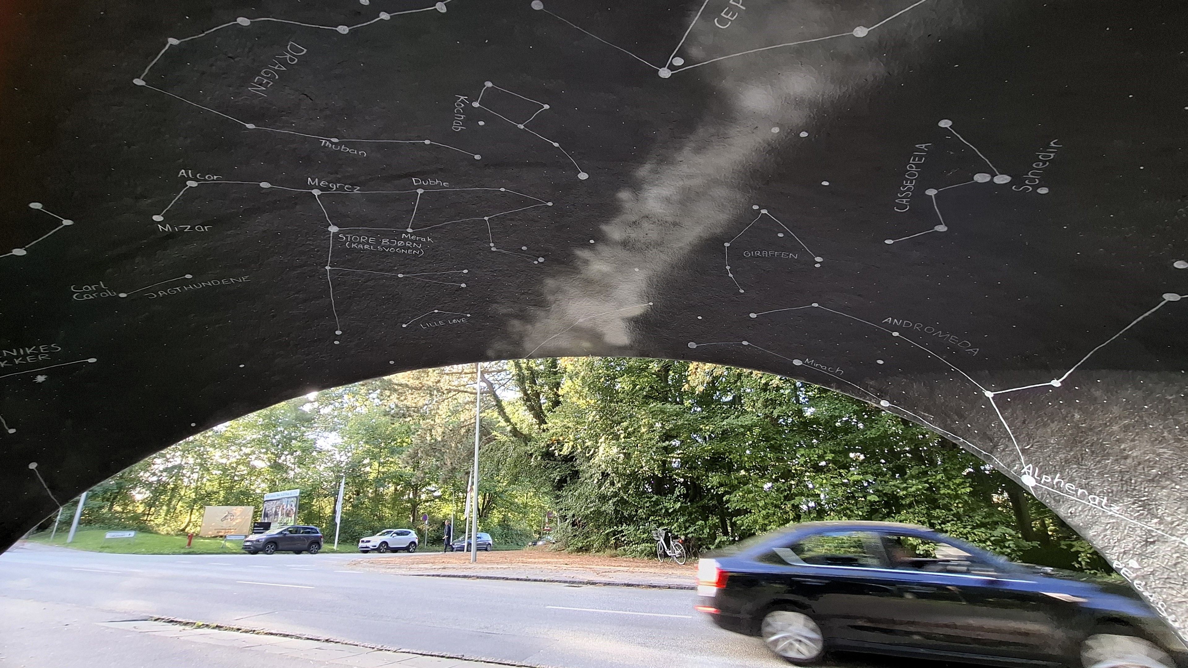 Inside the bus shelter looking out, a sliver of ‘night sky’ above our heads with stars and galaxies. Outside, a car passing by and other cars waiting to turn the corner. Now follows same full description as on all photos in the toot, so skip if you’ve already heard it. Aarhus. On the road outside the Ole Rømer Observatory, named after another famous Dane, Carl Nielsen. A broad clear road in Kongelunden, the Royal Woods, with trees on either side. Looking straight at the bus shelter, one of the city’s iconic mushroom-shaped bus shelters designed in the 1960s by furniture designer Poul Cadovius. A low concrete mushroom-shaped shell with standing room and a little bench inside. The council commissioned artists to paint them with local motifs.  Artist Ekaterina Poliakova painted the night sky in this one. As you wait for the bus, you are surrounded on three sides and above by a black night sky with a map of the stars and constellations and galaxies, their names written beside them.  Outside, the shelter’s shell is painted like a day sky – light blue with fluffy white clouds.
