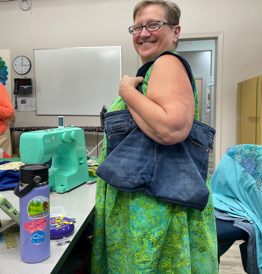 A woman holds a denim tote bag, sewn from a pair of jeans. A teal green sewing machine sits nearby.