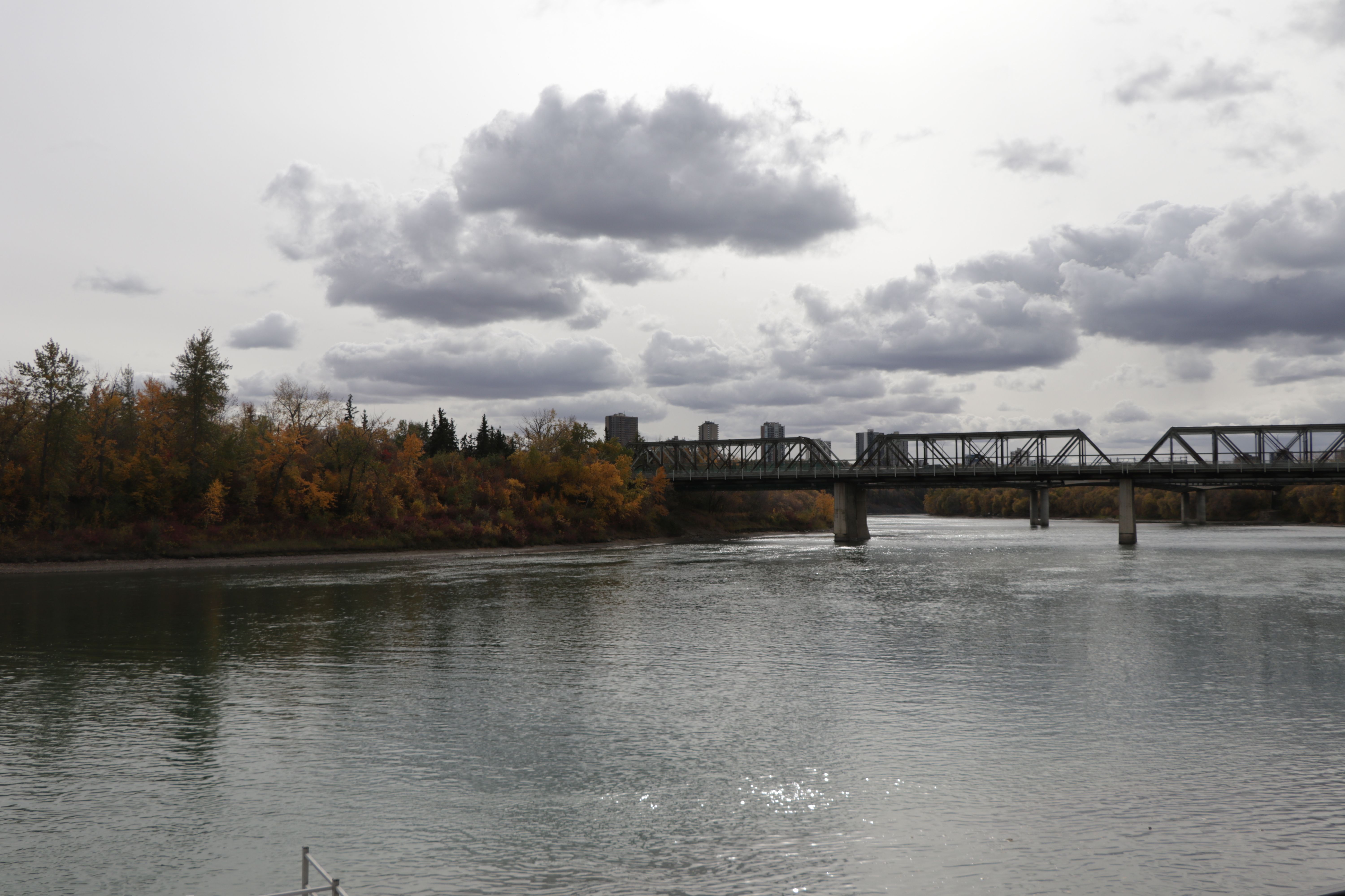 View near the edge of a shimmering river with silver lined clouds in the distance and a bridge. In the back you can see some taller apartment buildings.