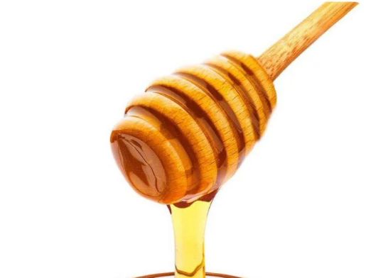 A close-up studio shot of a wooden honey dipper, coated in golden-brown honey, with a stream of viscous honey dripping down from the bottom against a clean white background.