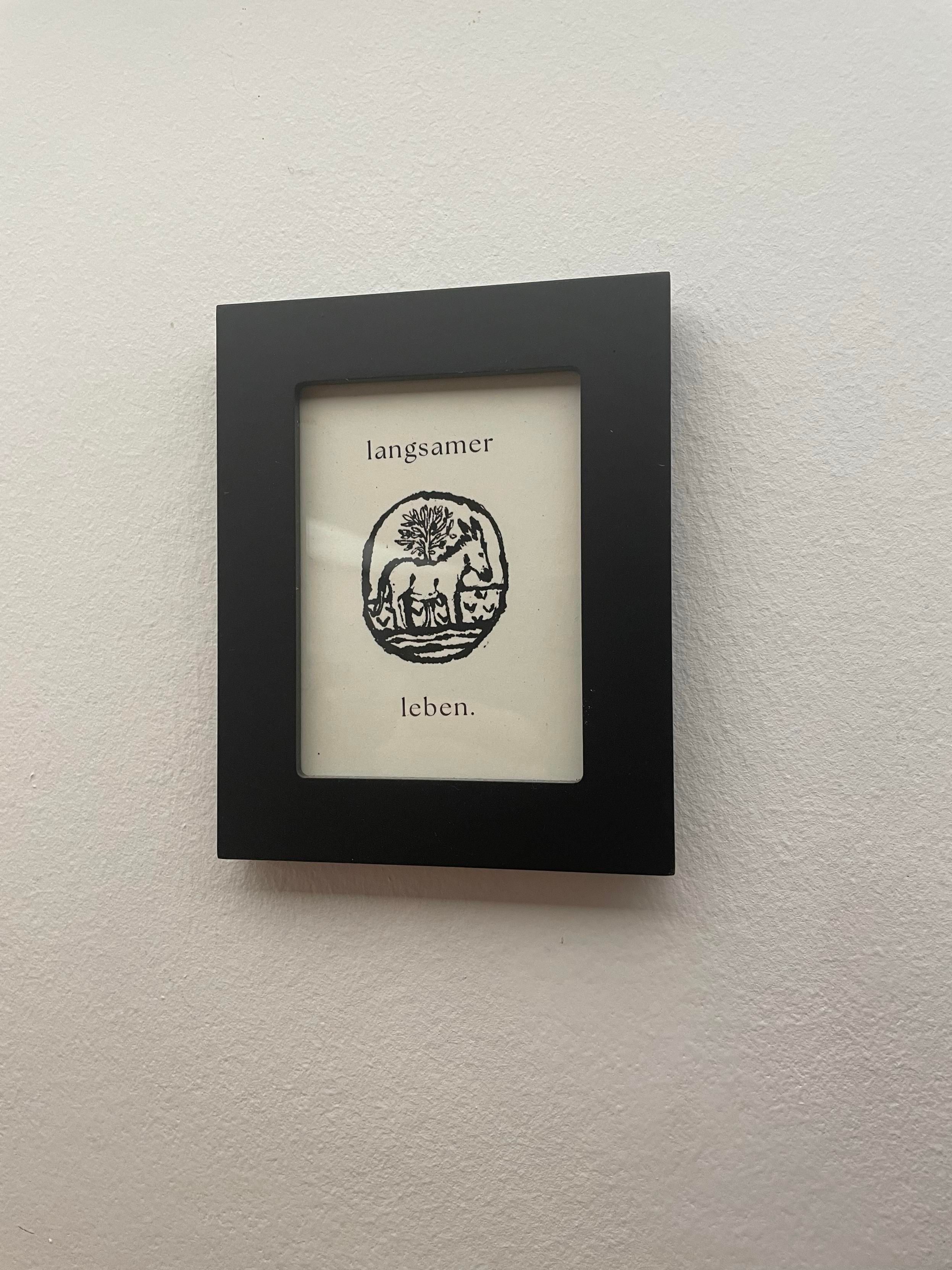 A framed artwork featuring the German phrase "langsamer leben" (meaning "live slower") above an illustration of a donkey with a tree design. The frame is black and the image is hung on a light-colored wall.