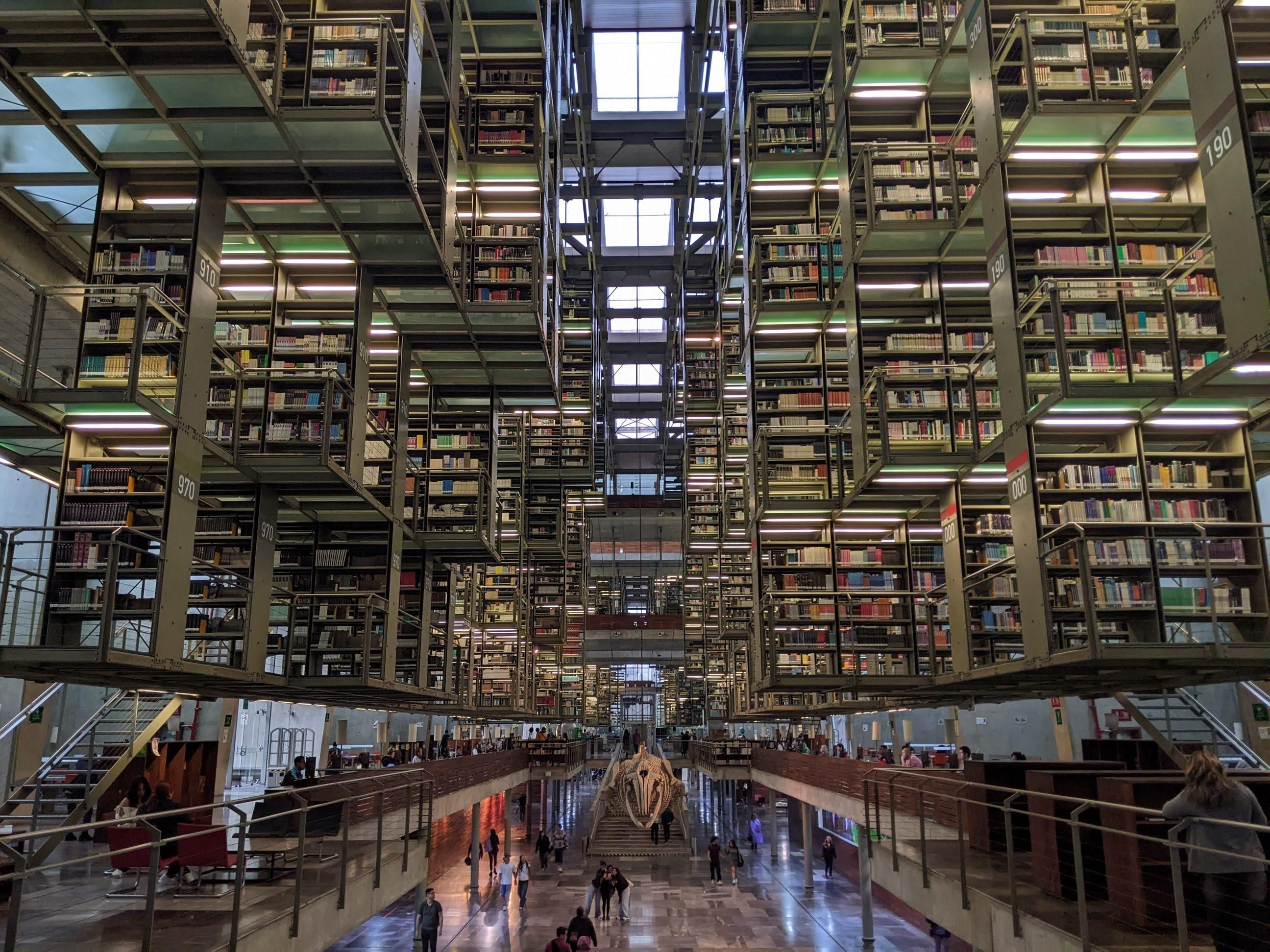 A look inside the Biblioteca Vasconcelos in Mexico City. There are columns of bookshelves seeming suspended at various levels from the ceiling with walkways connecting and that keep going into the far distance. Light from glass skylights many stories up and interior lighting are throughout. On the bottom floor, slightly lower than the camera vantage is the skeleton of a whale