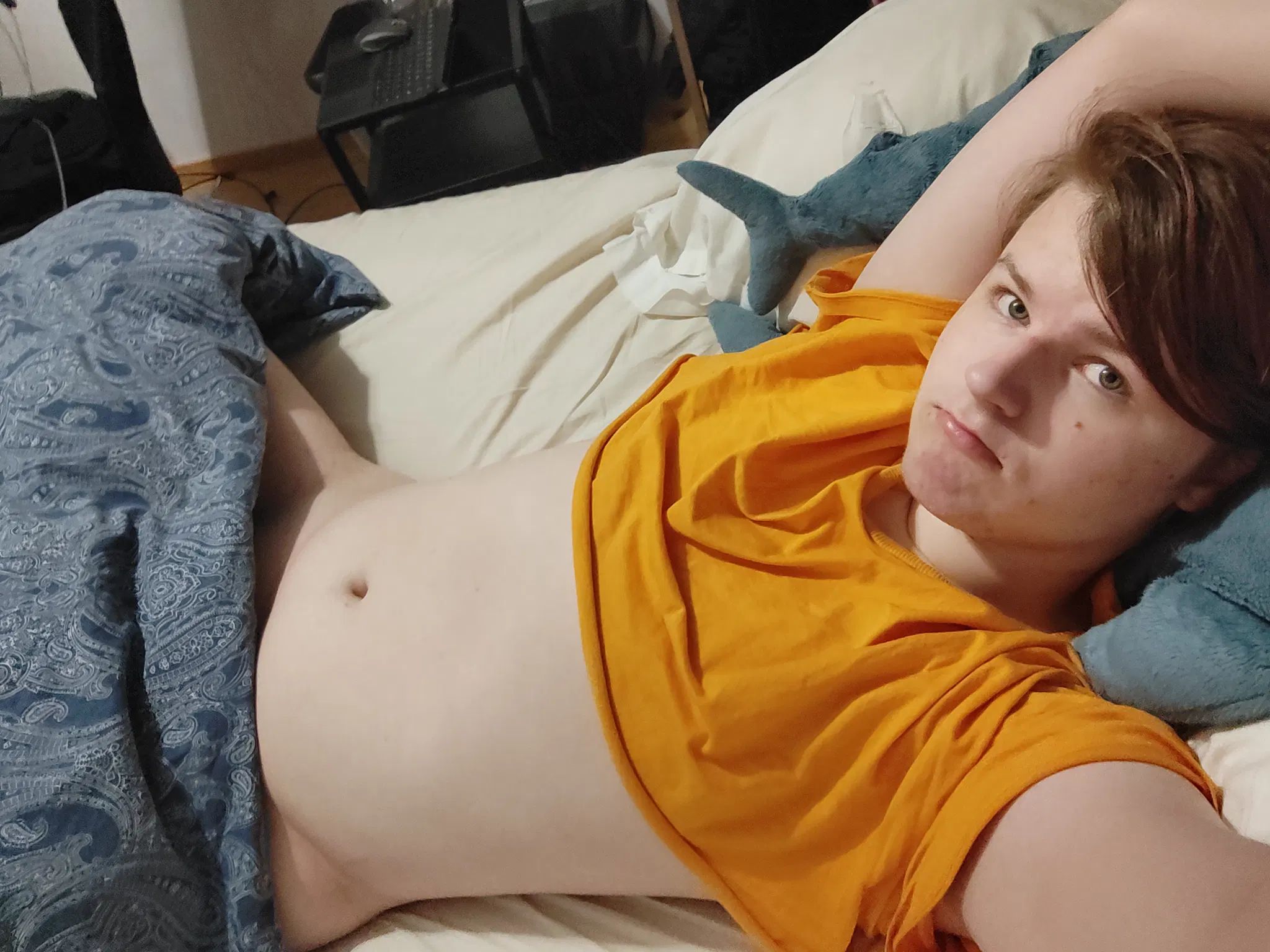 A teenager lying in bed. They are wearing an orange crop top and nothing else that we can see. A lot of tummy is shown. I'm the teenager. I'm resting my head on my BLÅHAJ and there is a baby BLÅHAJ near my head also. A blanket is covering my legs and crotch mostly, but you can see that I'm not wearing any underwear, my hips are visible, and a little bit of thigh. In the background, there is an office chair with some clothes on it and a metal nightstand with a laptop on it, as well as a mouse. I'm looking at the camera with a 🥺 expression, all adorable like. My hair is a mostly dark brown, faded purpleish color. My top is covering enough to not show my breasts, but I'll say this again: a lot of tummy is visible (and in fact it is the main focus almost)