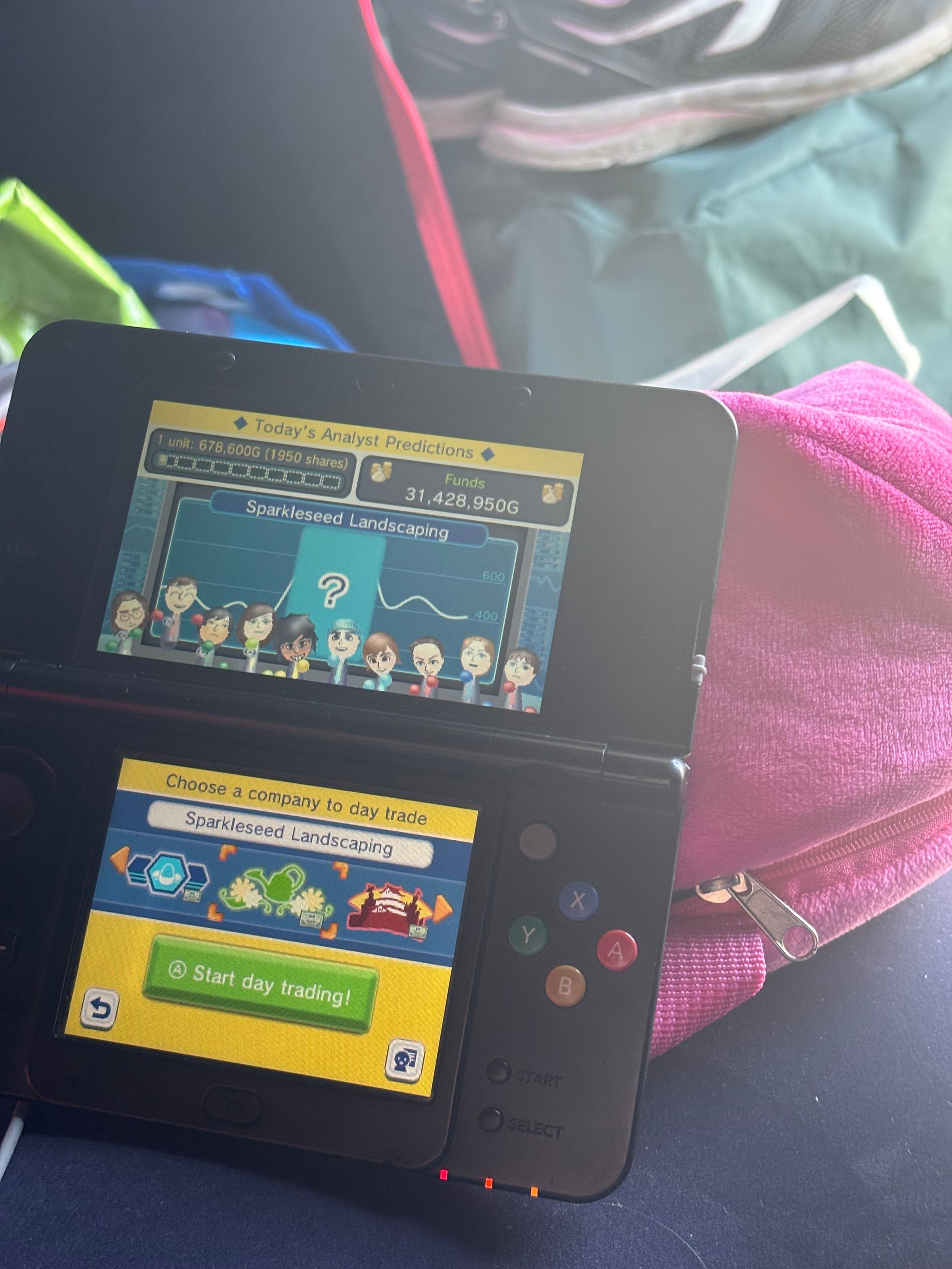 Photo in a tent of a black New Nintendo 3DS running StreetPass Trader, a game about fictional stock trading where the Miis of people you meet on StreetPass help you out as analysts. There are some bags in the background, including a pink octopus backpack.

Ten Miis are on the top screen, one of whom is jumping up and down excitedly about a company, Sparkleseed Landscaping, which is predicted to rise to maybe a big peak in the middle of trading on a chart.

On the bottom screen, a menu letting me scroll through companies, and button marked ‘Start day trading!’