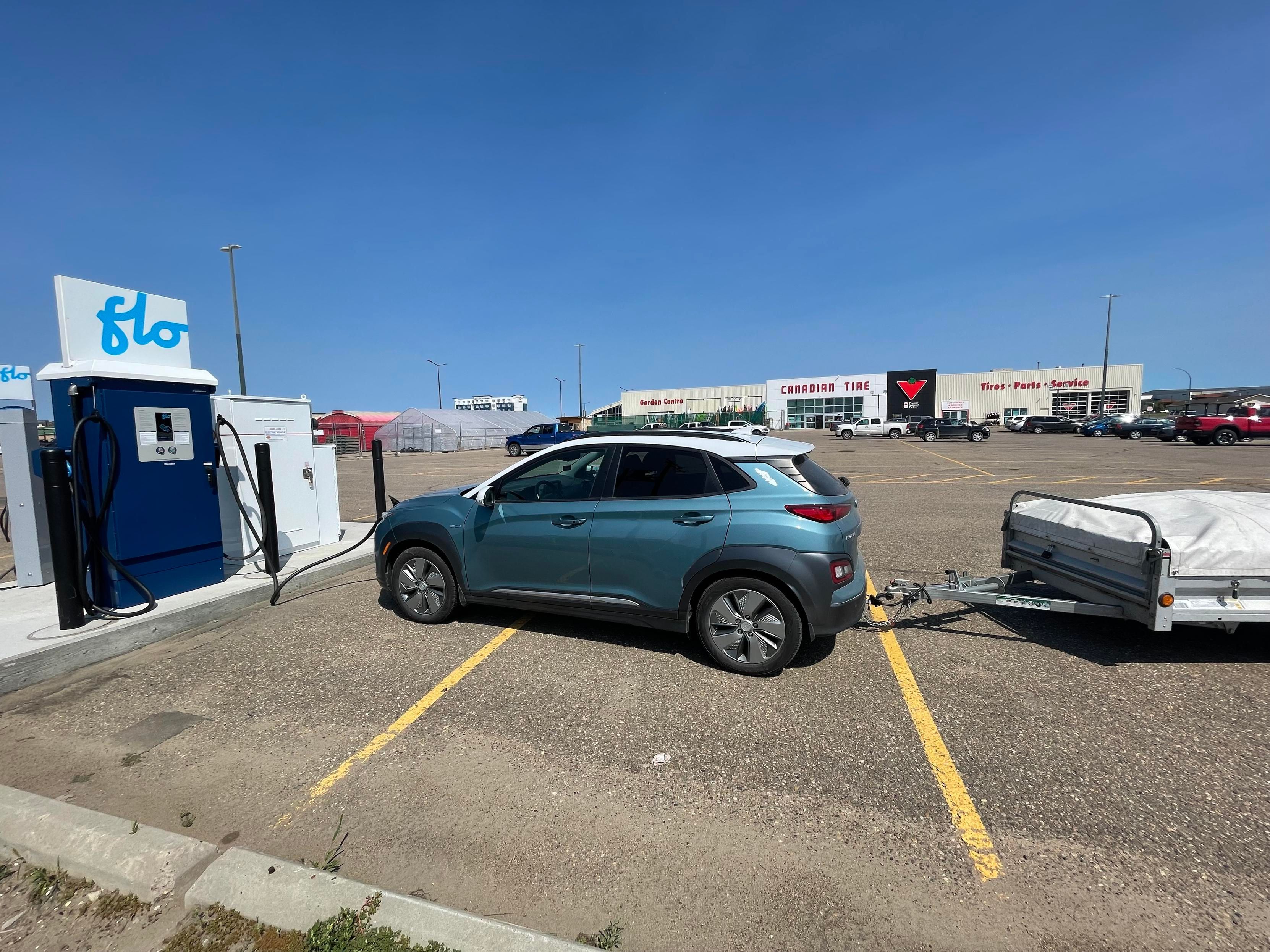 The turquoise coloured Kona car pulling a small trailer is pulled up to a Flo charger in a large parking lot. In the distance is Canadian Tire. The sky is big and blue.
