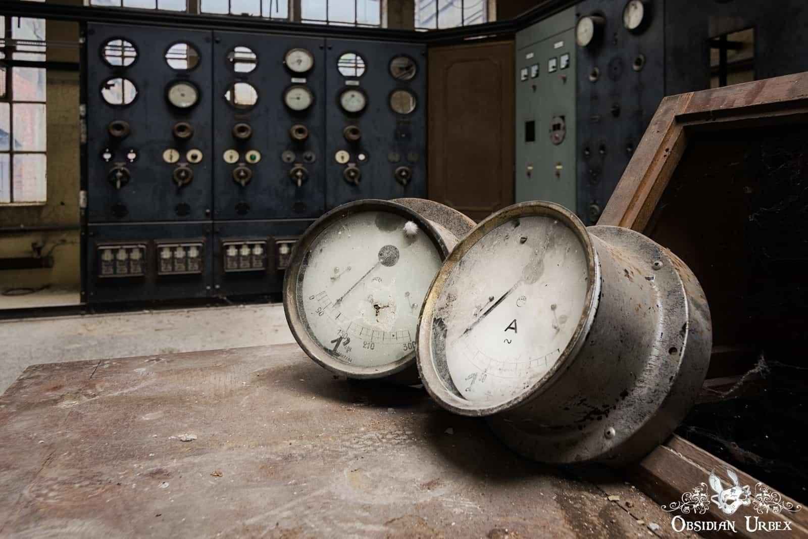 A photo of an abandoned industrial setting. Two large, dismounted gauges rest on a dirty surface in front of large control panels. 