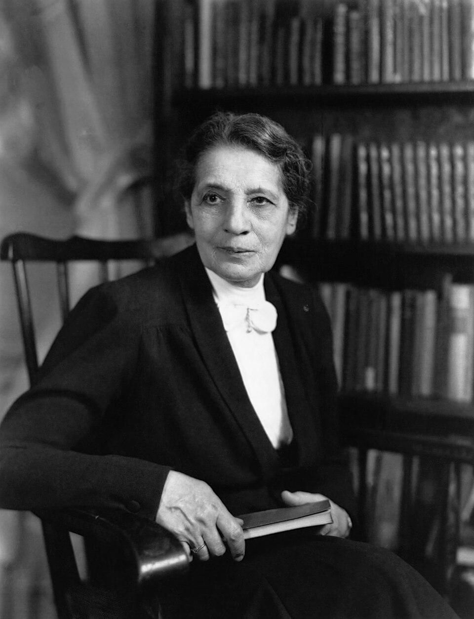 Dr. Lise Meitner, Austrian physicist, three-quarter length portrait.

A formal black and white portrait of Dr. Lise Meitner, the pioneering Austrian physicist, seated in professional attire with a book in hand before a library bookshelf. 

https://en.wikipedia.org/wiki/Lise_Meitner#/media/File:Lise_Meitner_NatGeo.jpg