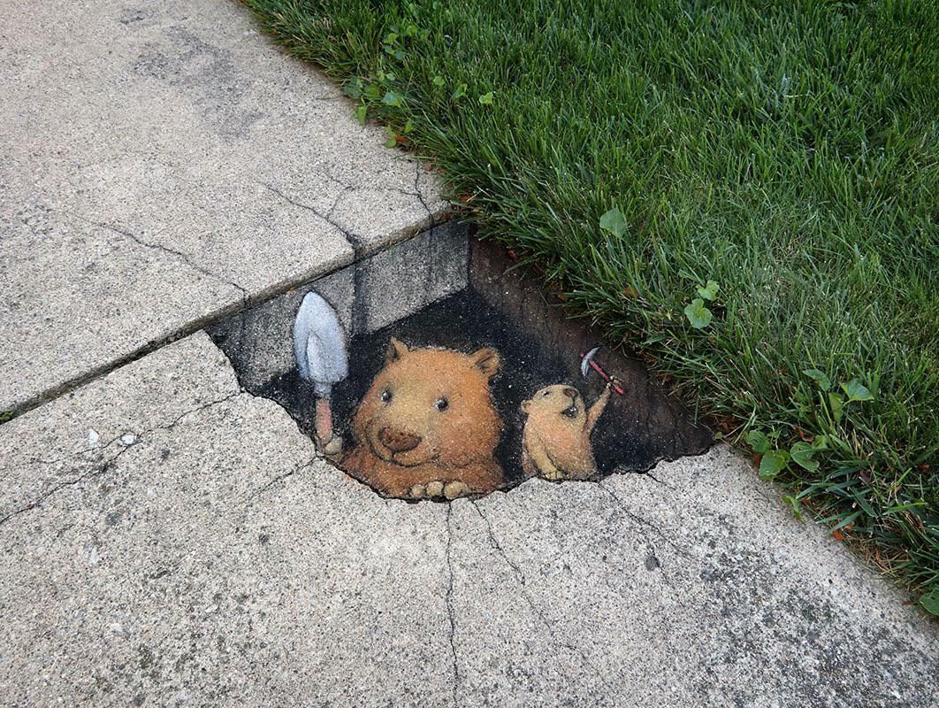 A chalk drawing of two small mouse like creatures, drawn on the pavement, emerging from a hole triumphantly as if they've dug their way to the surface. One carries a shovel, the other a pick
