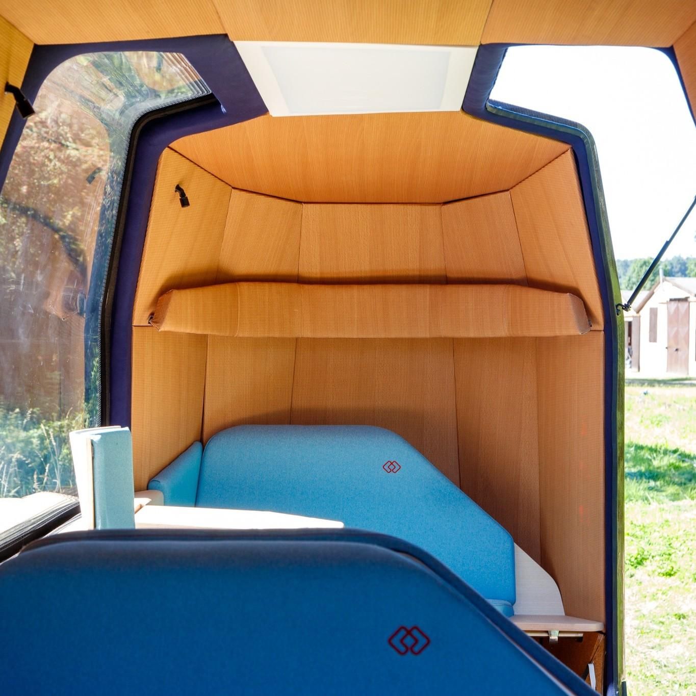 This image shows the interior of a compact, modern vehicle cabin with wooden paneling and large windows. The seating area features blue upholstered seats with a small table or surface nearby. The design is minimalistic and cozy, with natural light coming through the windows, revealing a grassy outdoor scene with a building and trees in the background. 