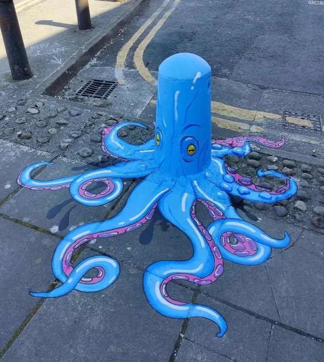 A bright blue octopus painted around a short street bollard in Limerick, Ireland, by Lumen Street Theatre. The cylindrical bollard forms the octopus’s head, complete with two cartoon-style yellow eyes and curved blue lines suggesting texture. The animal’s eight tentacles stretch out across the pavement in flowing pink-and-blue patterns, creating a playful illusion that the creature is spreading across the sidewalk and into the street. The artwork transforms an ordinary bollard into a lively marine character that appears to interact with the urban environment.