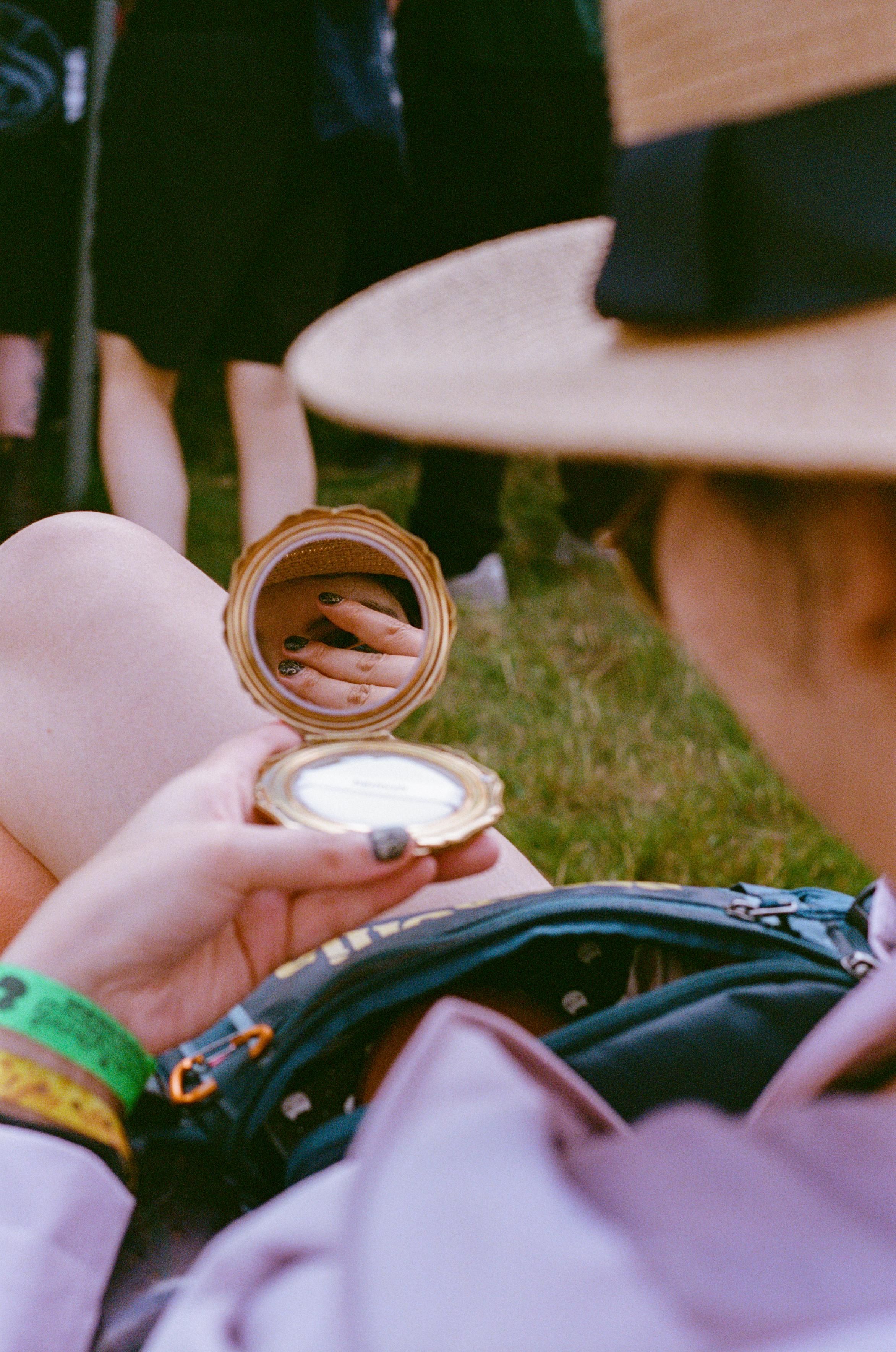 A woman wearing rain jacket and a straw hat is holding a tiny mirror in her hand while sitting on a camping chair on the grass. Through the mirror, a part of her face is visible, covered by her other hand.