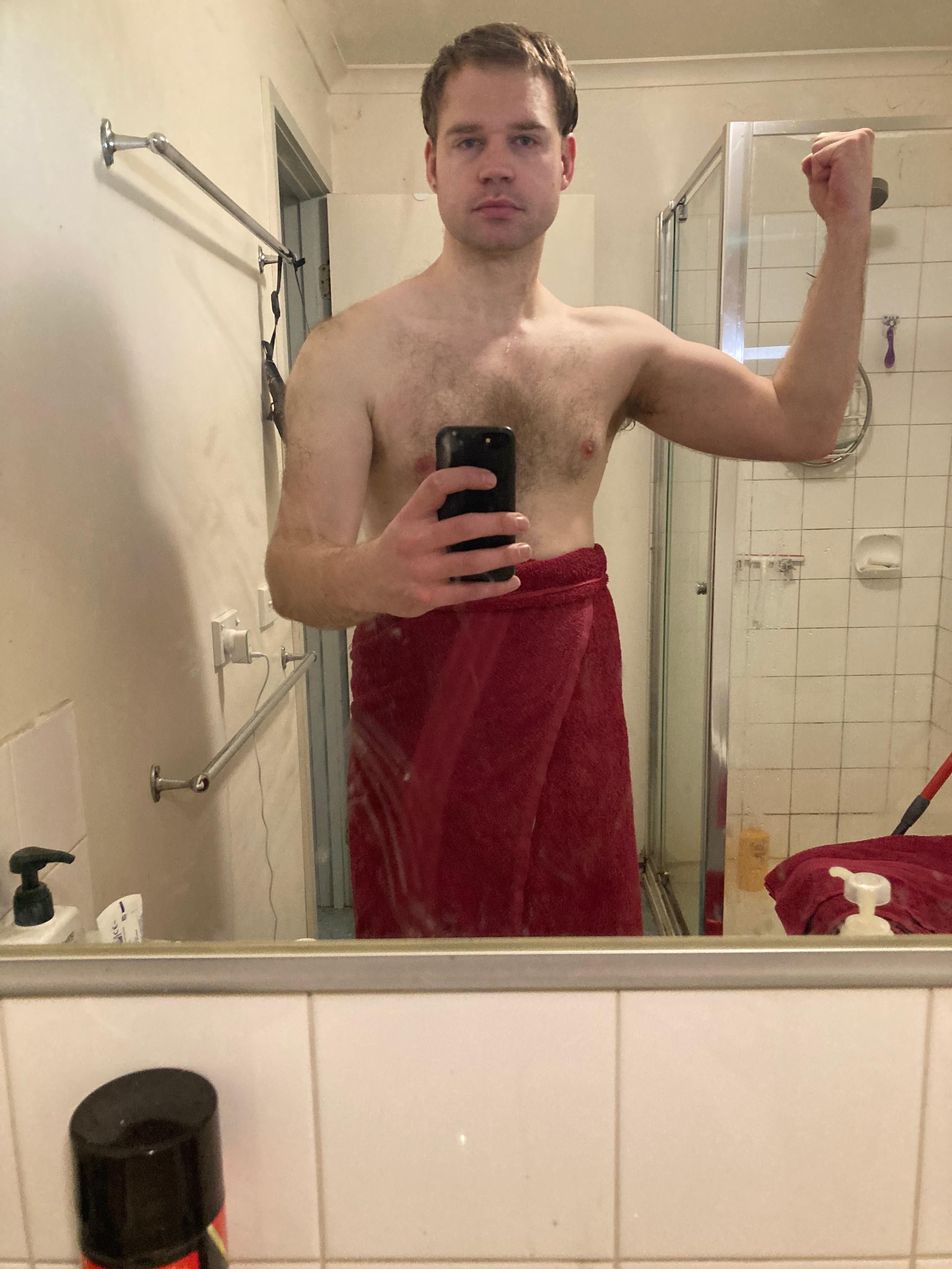 post-shower bathroom selfie of Luke. They are flexing their arm. They are wearing a red bath towel around their waist.