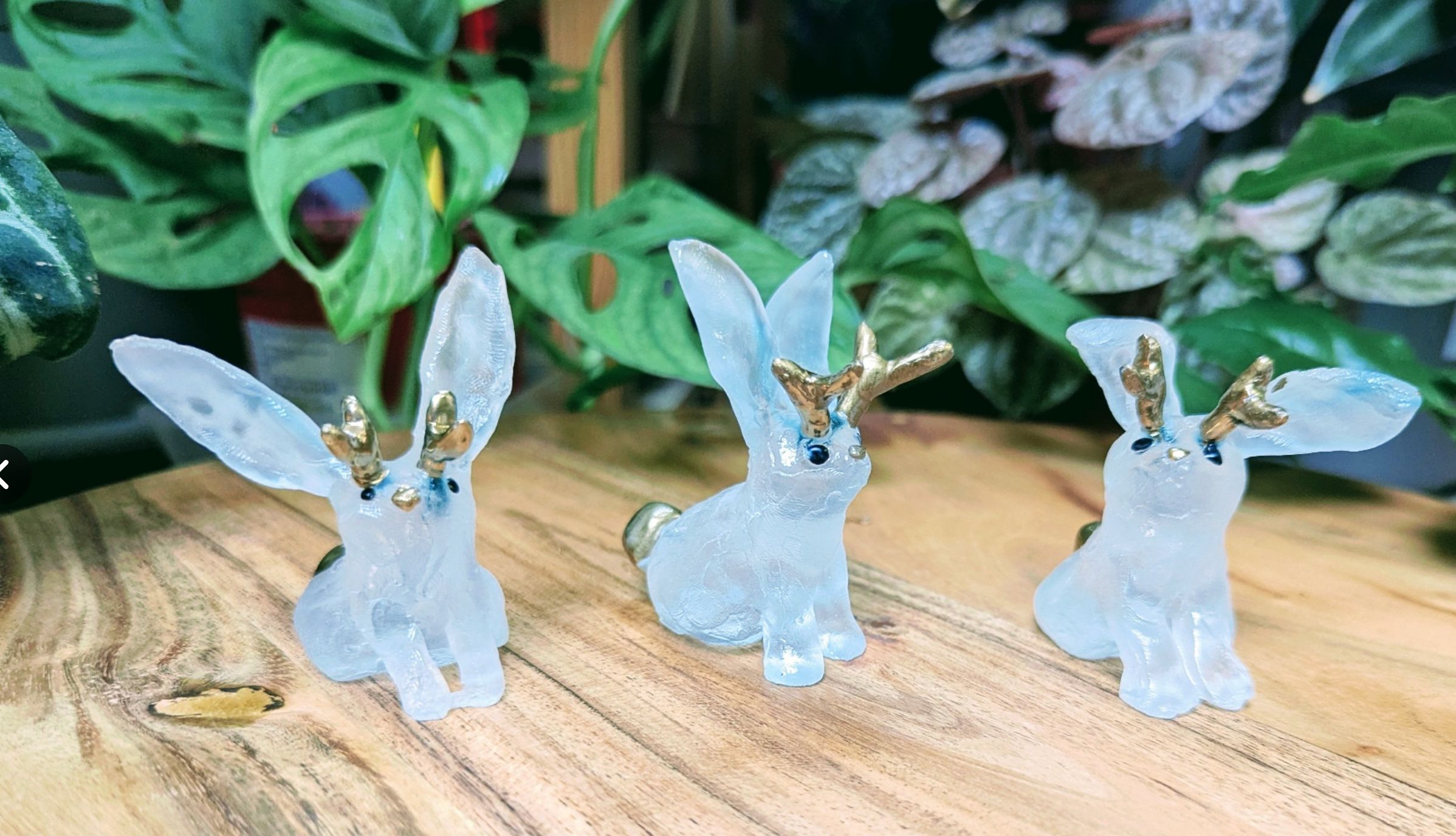 
Trois petits lapins en verre. Ils ont des petites cornes dorées en pompon doré ainsi qu'un petit nez doré. Ils sont posés dans un décor avec plein de plantes derrière