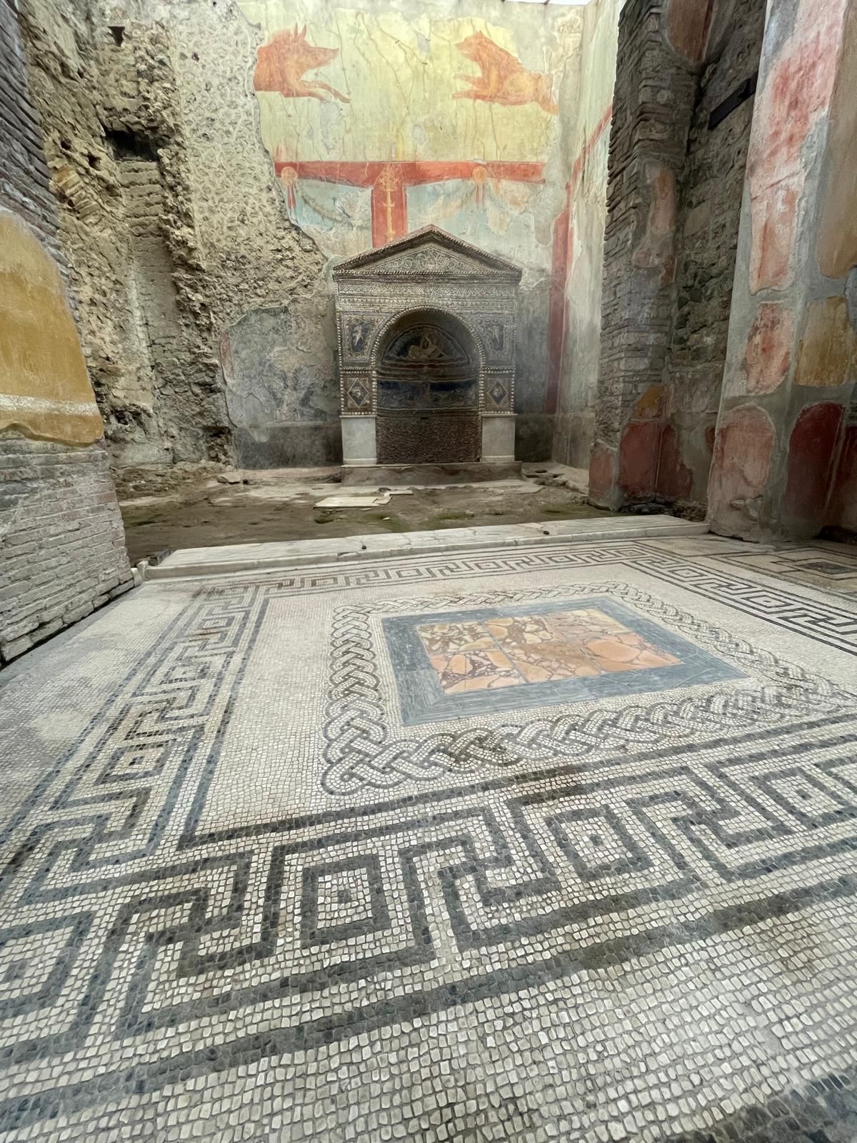 Interno della casa dell'orso ferito. Meraviglioso pavimento a mosaico geometrico. Pareti colorate. Sul fondo un'edicola interamente decorata con disegni a mosaico colorati. Sulla parete in alto, affresco raffigurante un cane a destra e il nostro amato cinghiale a sinistra che stanno per scontrarsi. Vincerà sicuro il cinghiale.