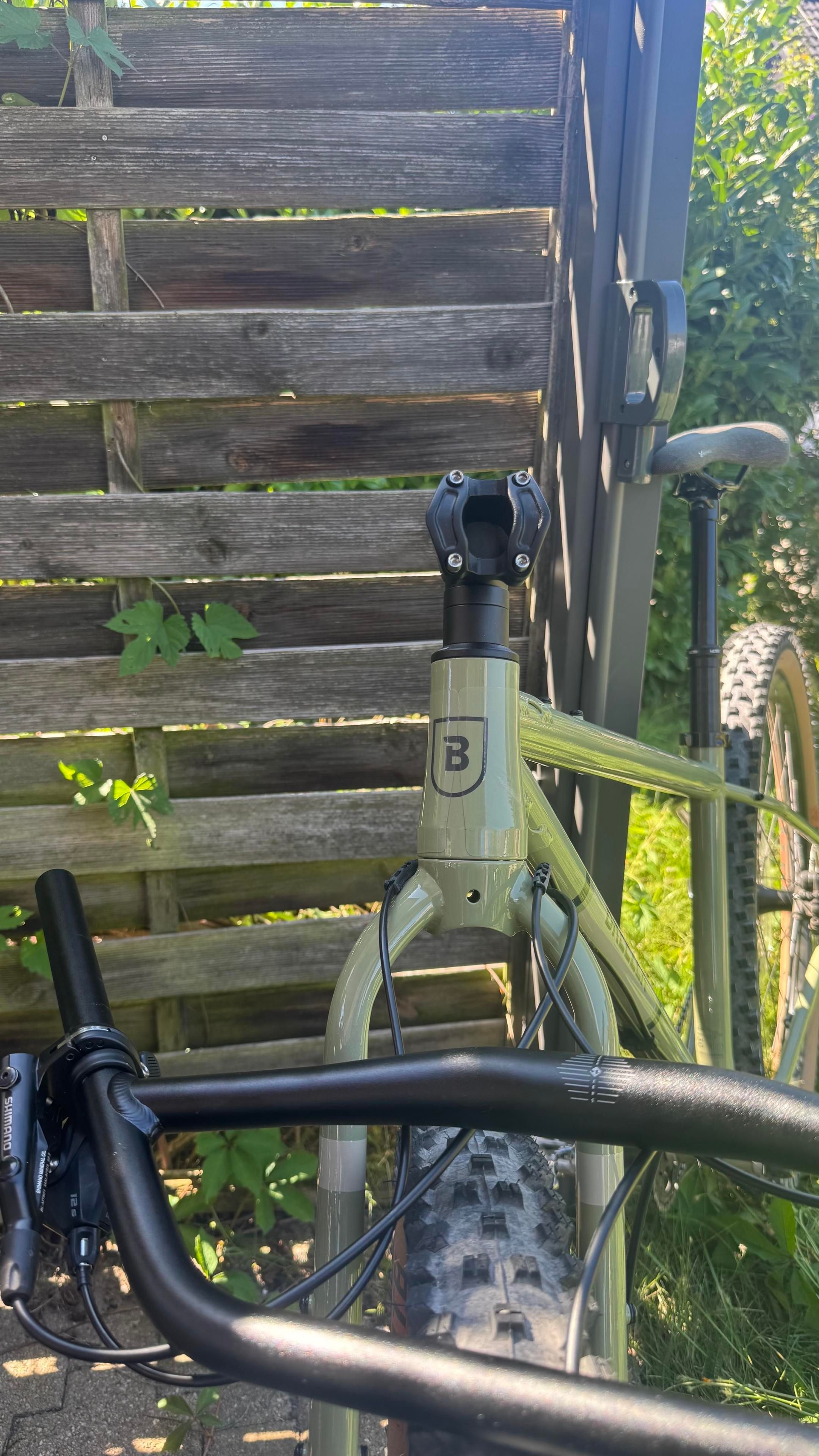 Eine Nahaufnahme des Lenkers und des Rahmens eines Fahrrads gegen einen Holzzaun. Das Fahrrad verfügt über ein leichtes Design mit einem markanten Logo auf dem Rahmen und einem gut getretenen Reifen im Hintergrund. Grünes Laub späht durch den Zaun
