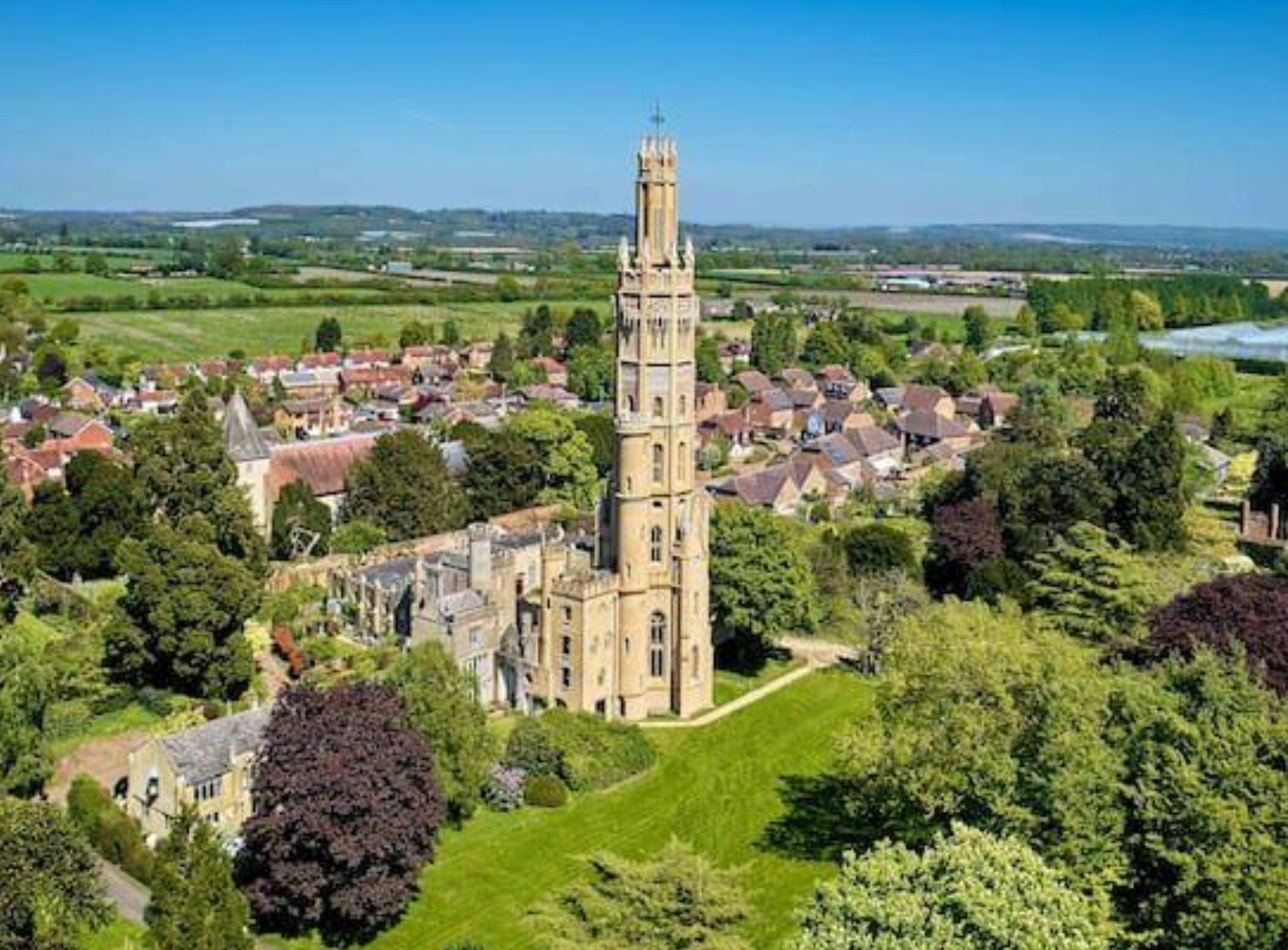 A gothic style stone tower that used to be part of a castle. it is 6 stories high and is surrounded by other small buildings and green landscape. 