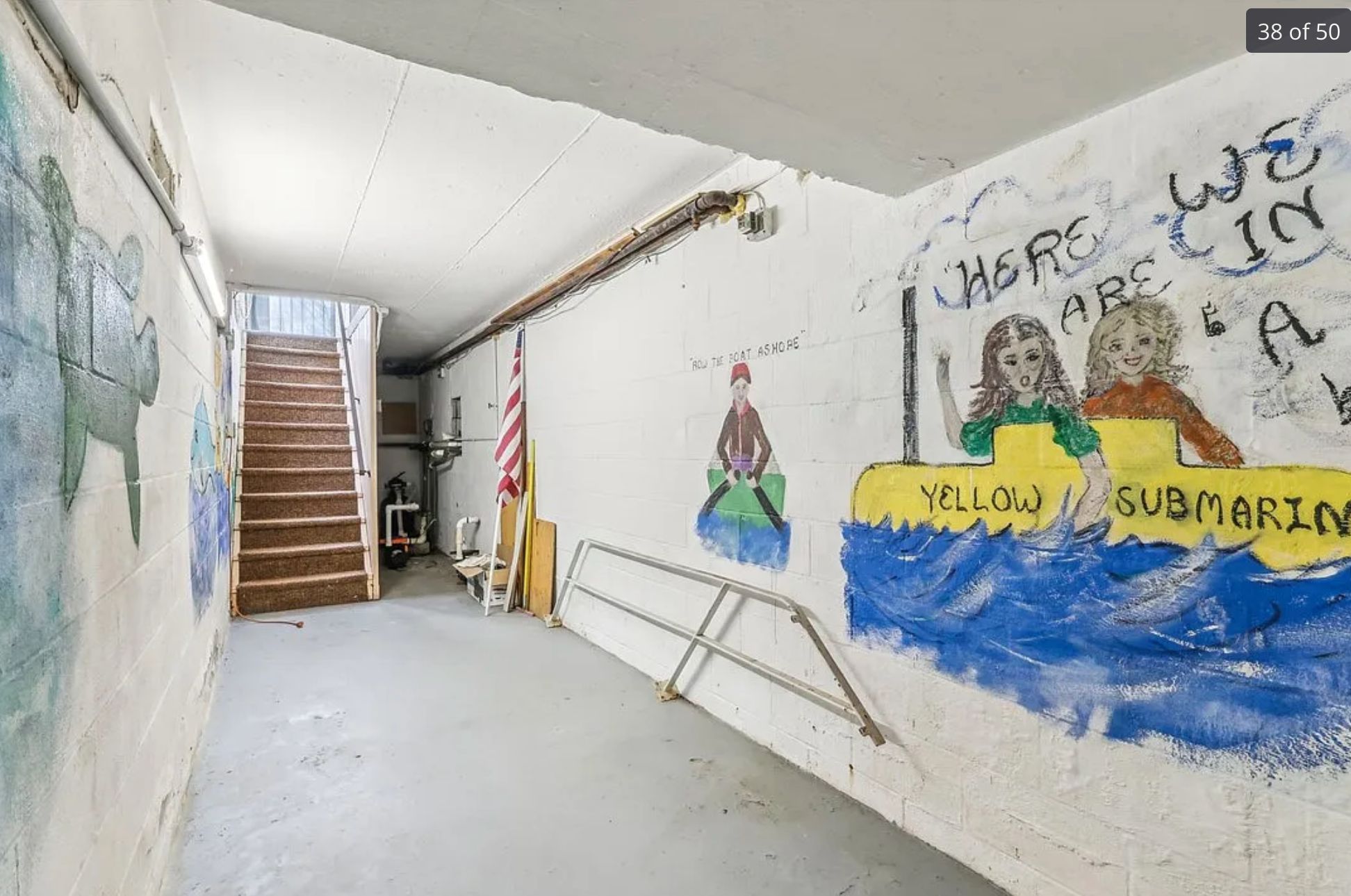 Another view of a room with a concrete floor and more crude murals, this one has a yellow submarine in water with text saying "we are in a", and it's assumed that off screen it says "yellow submarine". On the left wall there is a tortoise. The murals are worn and creepy. At the end of the room are stairs and an American flag propped up against the wall. 