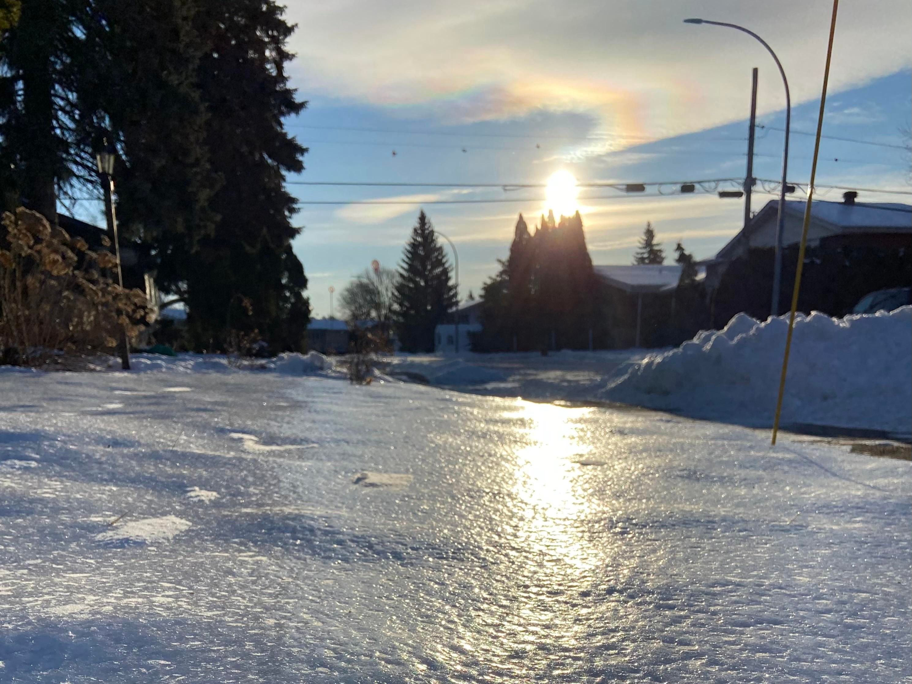 The sun is bright in the sky, just above some cedars and just below a strangely shaped cloud. The sun forms a rainbow over the cloud, like the top part of a question mark, with the sun itself being the door. The sun also reflects brightly in the shiny snowy yard in the foreground. 